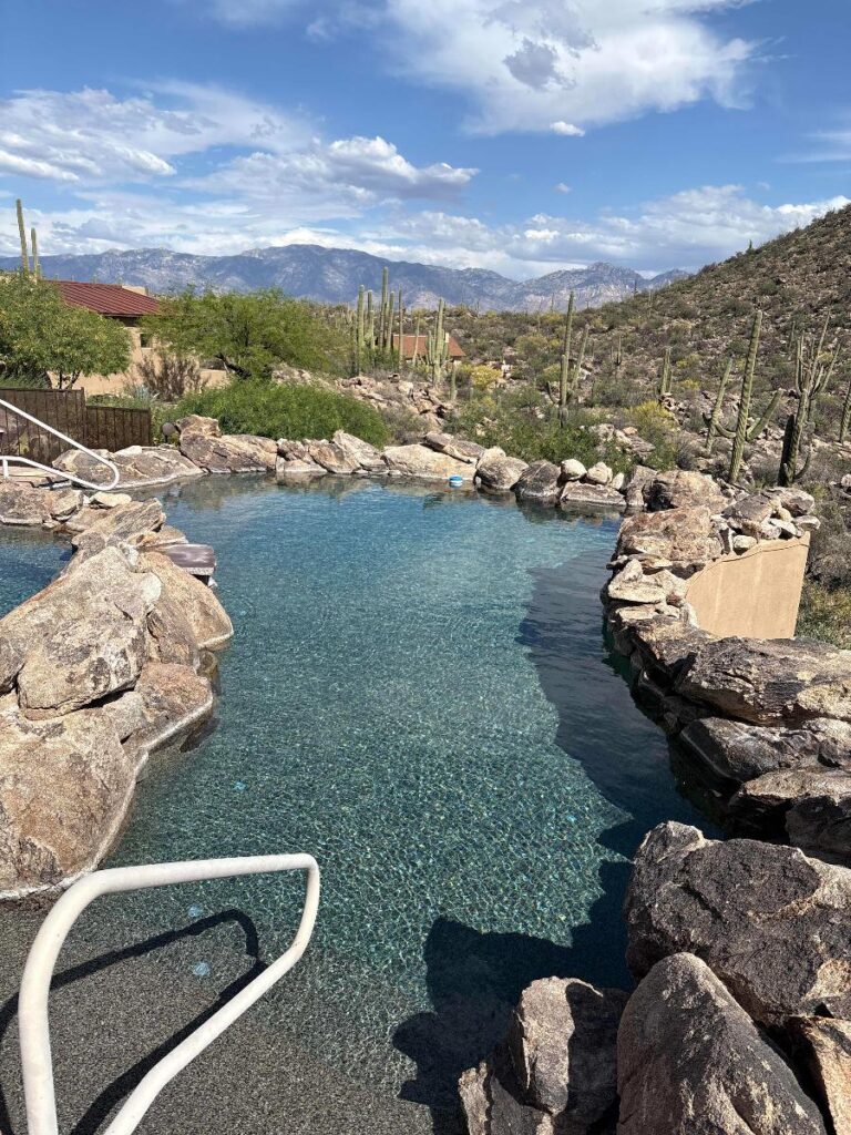 Rock-lined swimming pool in a desert landscape with cacti and distant mountains under a blue sky with clouds.