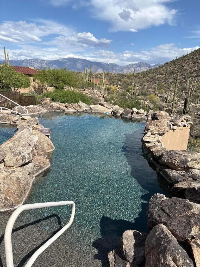 Rock-lined swimming pool in a desert landscape with cacti and distant mountains under a blue sky with clouds.
