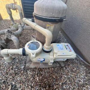 Outdoor pool pump and filter unit connected to PVC pipes along a gravel surface near a wall.