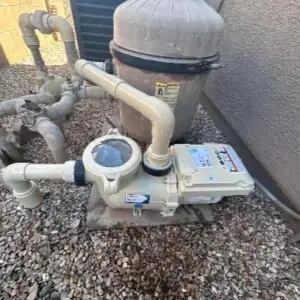 Outdoor pool pump and filter unit connected to PVC pipes along a gravel surface near a wall.