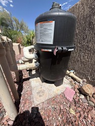 Above-ground pool sand filter with connected hoses, mounted on a stone patio beside a wall.