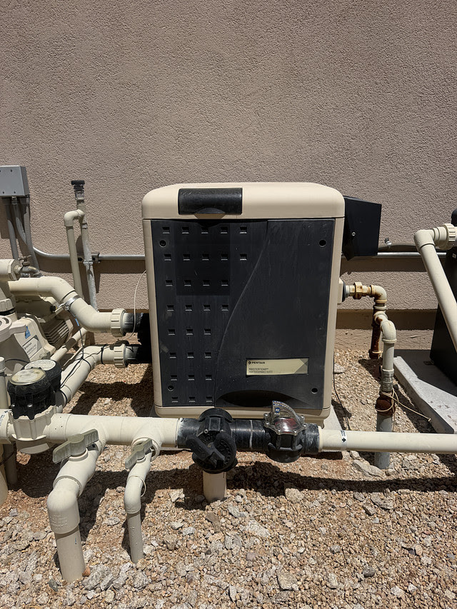 Outdoor pool equipment: a beige heat pump/filter unit connected to white PVC pipes and valves on a gravel surface near a stucco wall