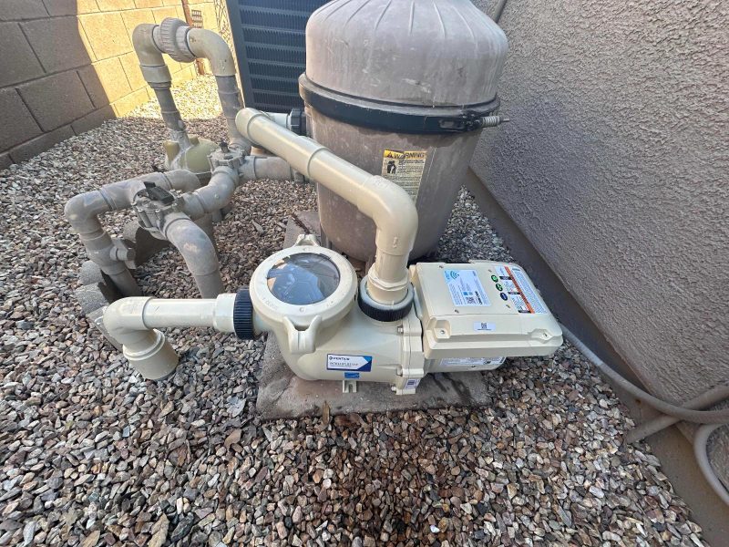 Outdoor pool filtration and pump system with PVC piping on gravel beside a stucco wall, including a round top-mounted filter canister and control box.