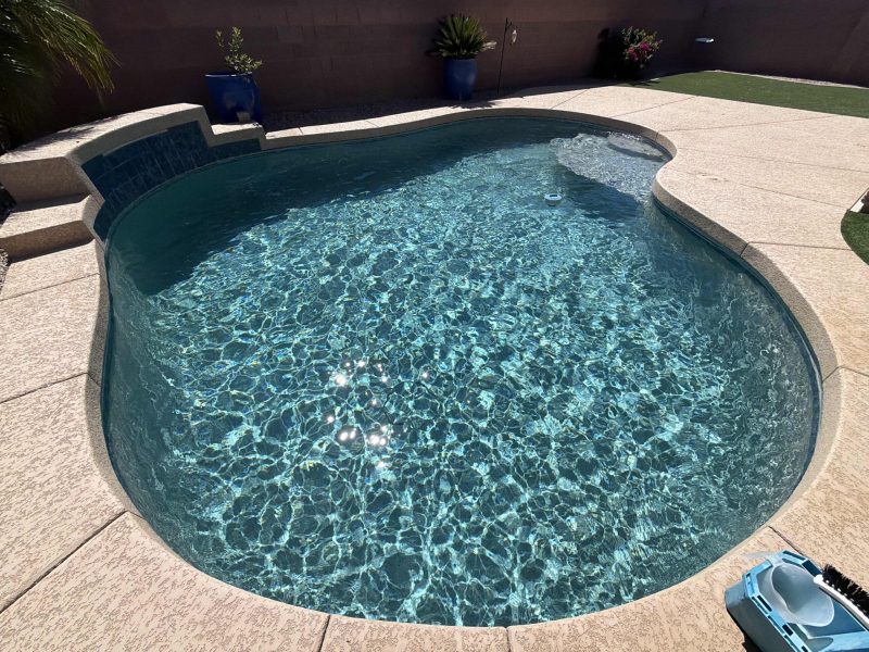 Curved in-ground swimming pool with clear blue water, sunlit ripples, and a beige textured deck surrounding it.